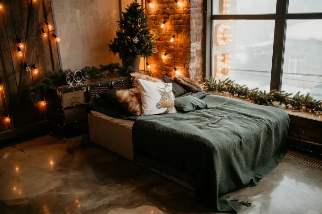اتاقی که تم آن زمستانی و کریسمسیه و پتو سبز روی تخت قرار دارد و ریسه ها به دیوار وصل شده اند و در کنار تخت پنجره ای به سوی طبیعت قرار دارد