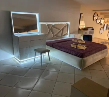 اتاق خوابی که دارای تخت و کمد و آیینه میباشد که دور تخت و آیینه و میز نور خورده است