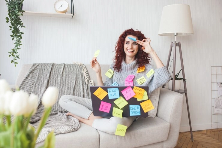 یک زن جوان و خوشحال و خندان با لپ تاپ و کاغذ یادداشت های رنگی نشسته بر روی مبل